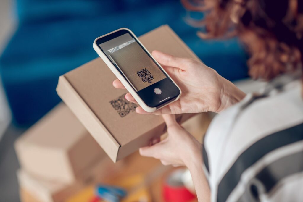 Cropped photo of a female worker aiming her camera at the QR code on the cardboard box. A metaphor of scanning a code for product passports.